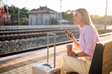 Cep telefonu kullanan, kahve içen, güneşli tren istasyonunda bir bankta oturan ve trenin gelişini bekleyen şık bir iş kadını..
