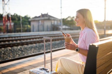 Şık bir iş kadını, güneşli tren istasyonunda bankta bavuluyla oturuyor, akıllı telefon kullanıyor ve trenin gelmesini beklerken kahve içiyor..