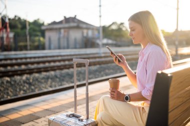 Kendine güvenen bir iş kadını, güneşli tren istasyonunda bankta bavullarıyla oturuyor, akıllı telefon kullanıyor ve trenin gelmesini beklerken kahve içiyor..