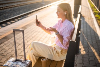 Güzel ve kendine güvenen bir iş kadını, güneşli tren istasyonunda bavullarıyla bankta otururken akıllı telefon ve kulaklık kullanıyor. Bir video görüşmesi sırasında konuşuyor ve el kol hareketi yapıyor..