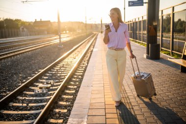 Zarif bir iş kadını bavuluyla güvenle yürüyor ve güneşli tren istasyonunda akıllı telefon kullanıyor. O, hareket halindeki modern banliyö ve bağlantıyı temsil ediyor..
