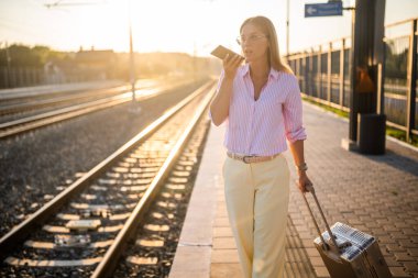 Modern ve kendinden emin iş kadını, güneşli tren istasyonunda bavuluyla yürürken cep telefonuyla konuşuyor..