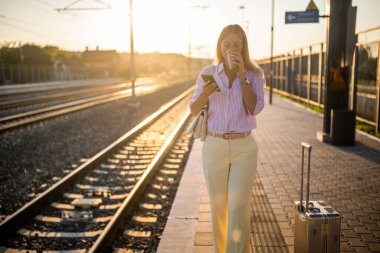 Şık bir iş kadını akıllı telefon kullanır ve kahve içerken güneşli tren istasyonunda elinde bavulla durup trenin gelişini bekler..