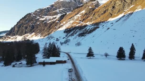 太陽の光の下で素晴らしい雪のスイスアルプスの山々の空中ビュー 観光客のためのいくつかの木造コテージと山の丘の麓にある小さなリゾート村 道路脇を通過駐車車 — ストック動画