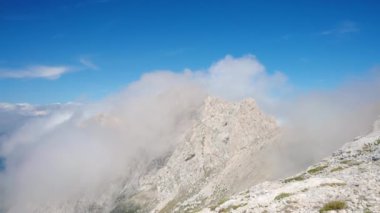 Puslu sis, açık mavi gökyüzü ve dağların tepesinden akan kalın dağlar ile kaplı beyaz kaya dağlarının nefes kesici panoramik görüntüsü, zirvenin zirvesinde rahatlama ve özgürlük hissi.