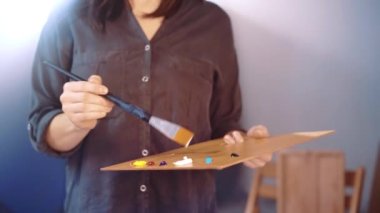 Talented artist against half illuminated workshop background holding wooden palette and mixing different oil colour with paintbrush, inspired painter preparing tools for painting on canvas in workshop