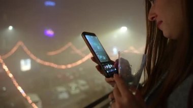 Trendy cellphone in hands of pretty smiling woman standing on balcony against amazing illuminated streets background, woman taking pictures or recording video of festive street illumination