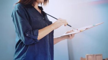 Female artist against illuminated blurred workshop background, woman mixing different oil colours on wooden palette with paintbrush, painter preparing tools for creating piece of art on canvas