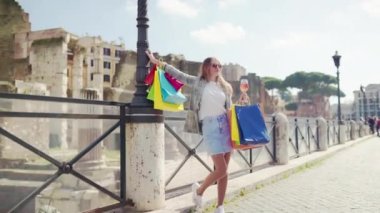 Glamour lady in sunglasses with shopping bags in the city centre against ancient Roman architecture background, stylish woman drinking aperol spritz cocktail while walking the street, female