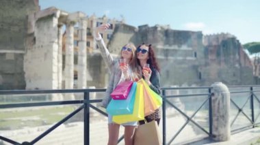 Stylish women in sunglasses taking selfie photos on amazing Roman architecture background, gorgeous ladies relaxing and enjoying their shopping day in the city centre, sexy women with shopping bags