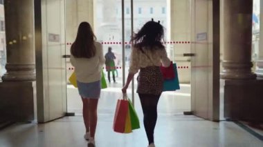 Back view shot of walking fashion ladies with shopping bags, happy women leaving big shopping mall centre with filled shopping packages, luxury lifestyle of elegant women, sales shopping concept. High