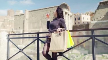 Charismatic woman with glass of refreshing cocktail in hand watching around and admiring ancient Roman architecture, female tourist relaxing and enjoying outdoor after shopping in fashion boutiques