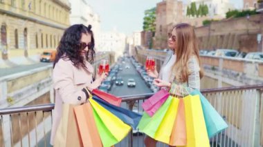 Female friends meeting in the city centre and drinking refreshing aperol spritz cocktail after perfect shopping day, smiling ladies talking and flirting on camera, stylish women with shopping bags in