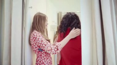 Female friends doing shopping in clothes store, ladies trying on outfit assortment searching for suitable wear, women in fitting room choosing glamour outfit, shopaholic concept. High quality 4k