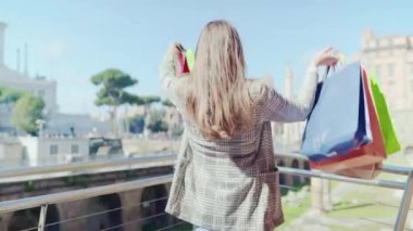 Happy woman against landmark background lifting hands up with colourful shopping bags, stylish female enjoying her purchases in the city centre, joy and happiness of young woman doing shopping with