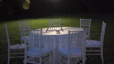 Round table covered with tablecloth and white chairs in the garden at night, small candle burning in the centre of the table, spot light on background illuminating table setting, empty garden