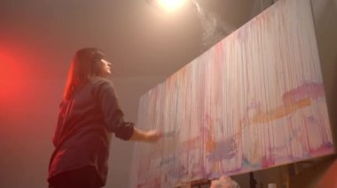 Professional female artist creating contemporary masterpiece in dark illuminated workshop, painter spraying canvas with watercolours drops using paint brushes, innovation in artistic oil painting