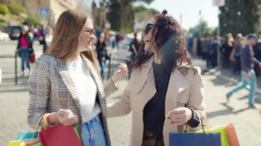 Female friends walking across the city centre street with colourful shopping bags, trendy females relaxing outdoor after great shopping day in the mall and fashion boutiques, attractive women enjoying