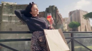 Italian fashion model posing and flirting on camera in the downtown of ancient Rome, stylish woman holding shopping bags and refreshing cocktail in hands, female touching her hair in blowing wind