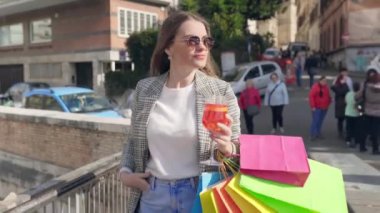 Fashion lady in sunglasses with shopping bags walking in the city centre, stylish woman drinking aperol spritz cocktail while walking the street, female shopaholic relaxing and enjoying shopping day