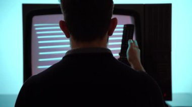 Problem of loneliness and TV addiction, back view shot of concentrated man sitting in front of television screen, watching broadcasting programs and changing channels with remote control, bad