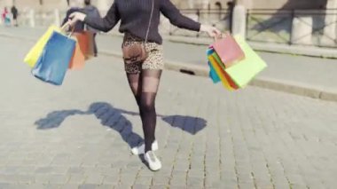 Stylish female tourist with colourful shopping bags walking across the streets of ancient Roman city in daylight, smiling shopaholic flirting on camera enjoying weekend relax and new outfit purchases