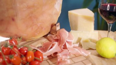 Ahşap kesim tahtasında ünlü parma jambonu dilimleri, parmesan peyniri, kırmızı şarap ve domates ile servis edilen geleneksel kavrulmuş jamon bacağı, yüksek kaliteli İtalyan yemeği seçimi, et antipasto konsepti.
