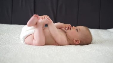 6 aylık bebek bezi içinde beyaz bir battaniyeye uzanmış, parmaklarını emip oynatıyor.