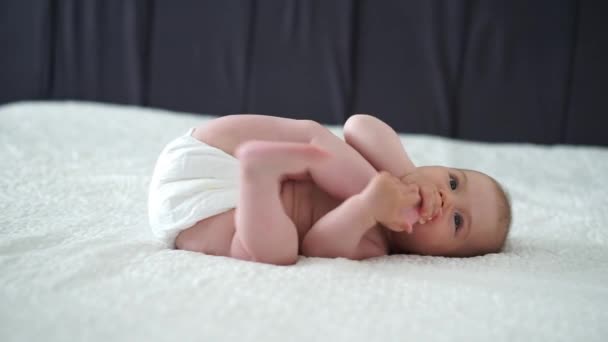 赤ん坊の女の子は白いシートに横たわり 足を握っています — ストック動画