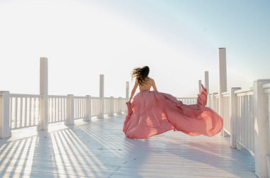 Güzel bir kız beyaz bir köprüde uzun pembe bir elbiseyle koşuyor.