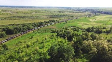 Uzun yük trenleri at arabalarıyla çorak tepe manzarasının yanında bir lokomotif taşır. Hava aracı bakış açısı yaz günbatımında. Dizel lokomotifli uzun bir yük treni..
