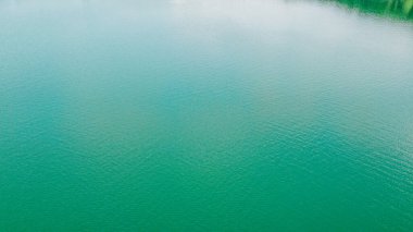 Aerial top-down flight over the amazing small lake of a perfectly round shape. Cloudy sky reflected in the clear turquoise water of a pond surrounded by trees and plants.