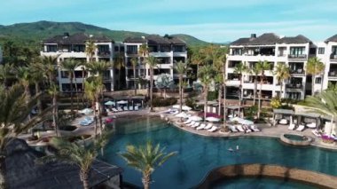 Zarif şemsiye ve sandalyelerin palmiye ağaçlarıyla çevrili olduğu tropikal bir tatil köyünde lüks havuz manzarası. Sakin atmosfer çarpıcı bir günbatımı ile güçlenir, Meksika, Cabo San Lucas 08.10.2024