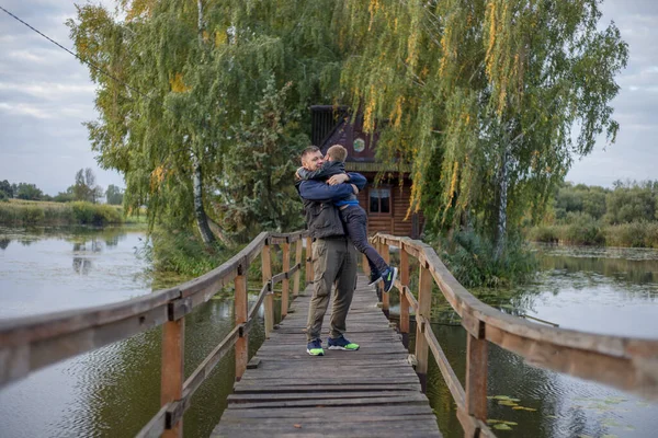 Happy father and son hugging and playing together in green nature. Fisherman\'s house with a wooden pedestrian bridge on a tiny island in the middle of the lake.