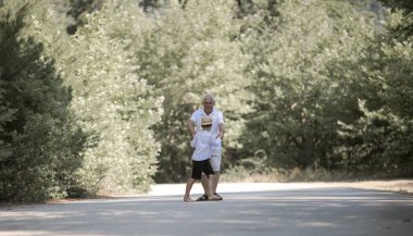 Büyükbaba ve torununun arka planda doğa yolunda yürüyen şapkaları var.