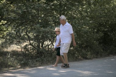 Büyükbaba ve torununun arka planda doğa yolunda yürüyen şapkaları var.