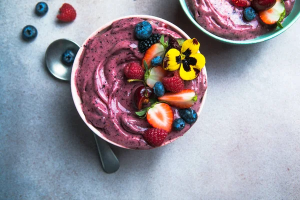 Two summer acai smoothie bowls with strawberries, blueberries,   on gray concrete background. Breakfast bowl with fruit and cereal, close-up, top view, healthy food