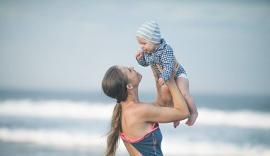 Neşeli genç anne ve küçük oğlu deniz kenarında birlikte eğleniyorlar, baba çocuğu havaya kusuyor.