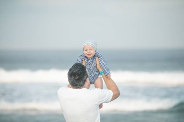 Neşeli genç baba ve küçük oğlu deniz kenarında birlikte eğleniyorlar, baba çocuğu havaya kusuyor.