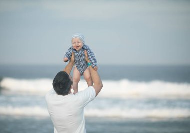 Neşeli genç baba ve küçük oğlu deniz kenarında birlikte eğleniyorlar, baba çocuğu havaya kusuyor.