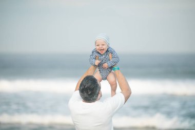 Neşeli genç baba ve küçük oğlu deniz kenarında birlikte eğleniyorlar, baba çocuğu havaya kusuyor.