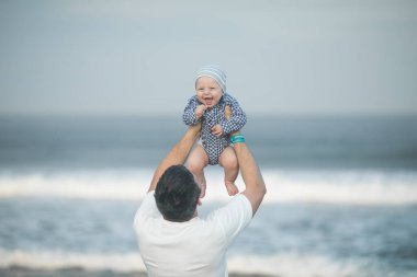 Neşeli genç baba ve küçük oğlu deniz kenarında birlikte eğleniyorlar, baba çocuğu havaya kusuyor.