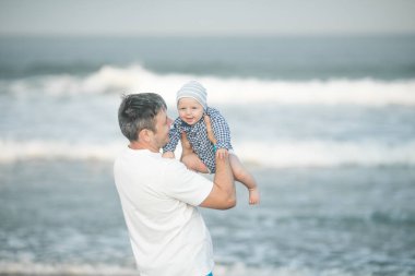 Neşeli genç baba ve küçük oğlu deniz kenarında birlikte eğleniyorlar, baba çocuğu havaya kusuyor.