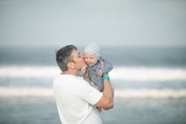 Neşeli genç baba ve küçük oğlu deniz kenarında birlikte eğleniyorlar, baba çocuğu havaya kusuyor.