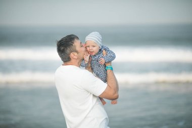 Neşeli genç baba ve küçük oğlu deniz kenarında birlikte eğleniyorlar, baba çocuğu havaya kusuyor.