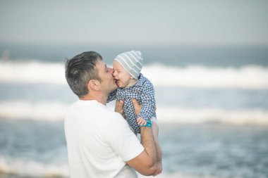 Neşeli genç baba ve küçük oğlu deniz kenarında birlikte eğleniyorlar, baba çocuğu havaya kusuyor.