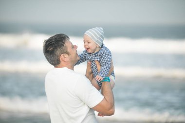 Neşeli genç baba ve küçük oğlu deniz kenarında birlikte eğleniyorlar, baba çocuğu havaya kusuyor.