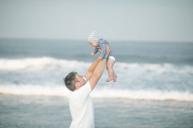 Neşeli genç baba ve küçük oğlu deniz kenarında birlikte eğleniyorlar, baba çocuğu havaya kusuyor.