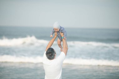 Neşeli genç baba ve küçük oğlu deniz kenarında birlikte eğleniyorlar, baba çocuğu havaya kusuyor.