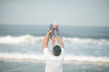 Neşeli genç baba ve küçük oğlu deniz kenarında birlikte eğleniyorlar, baba çocuğu havaya kusuyor.